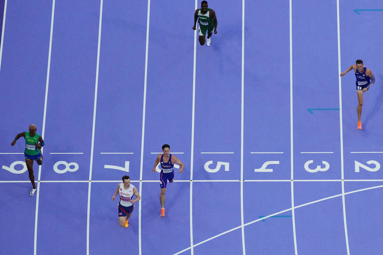 A imagem mostra uma corrida em uma pista de atletismo azul. Há cinco corredores visíveis, com dois deles em destaque na parte inferior da imagem, enquanto os outros três estão mais atrás. Os corredores estão usando uniformes de diferentes cores, e a pista possui marcas numéricas indicando as raias. A perspectiva é de cima, mostrando a dinâmica da corrida.
