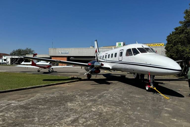 A imagem mostra um avião de pequeno porte branco com detalhes em preto e vermelho, estacionado em uma pista de aeroporto. Ao fundo, há um hangar e outro pequeno avião. O céu está limpo e azul, e a grama aparada ao lado da pista 