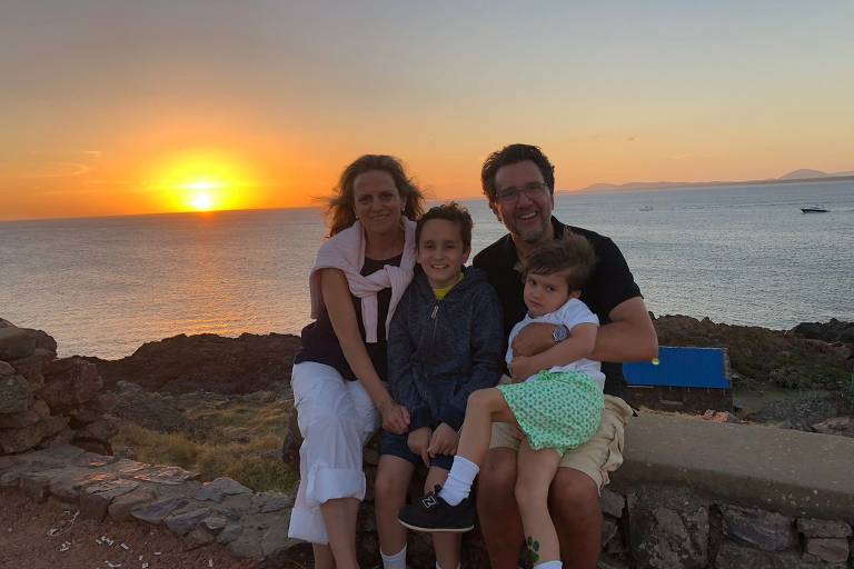 Uma família sentada em uma pedra, com o mar ao fundo e um pôr do sol laranja. A mãe está à esquerda, usando uma blusa escura e calças brancas, com um suéter amarrado nos ombros. O pai está à direita, segurando uma criança pequena. Entre eles, um menino está sentado. O cenário inclui rochas e um céu claro.