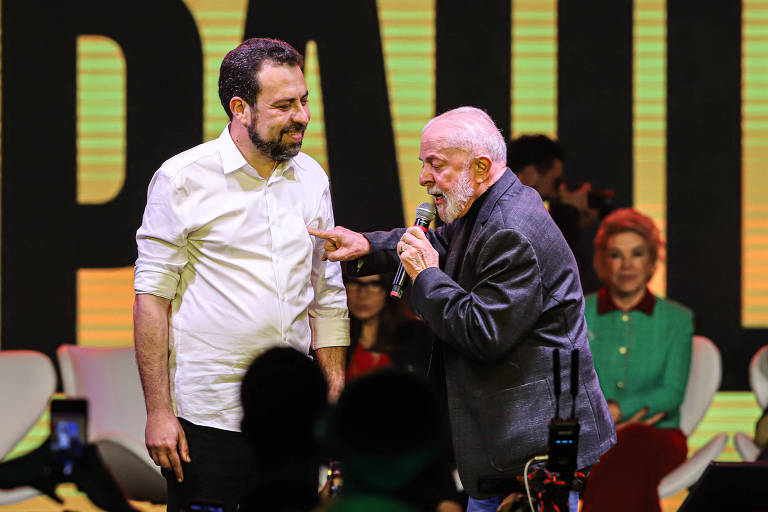 A imagem mostra duas pessoas em um palco durante um evento. Uma delas, vestindo uma camisa branca, está em pé ao lado de um homem mais velho, que usa um paletó escuro e está falando ao microfone. Ao fundo, há uma tela iluminada com o nome 'PAUL' e outras pessoas sentadas em cadeiras, algumas com roupas coloridas.