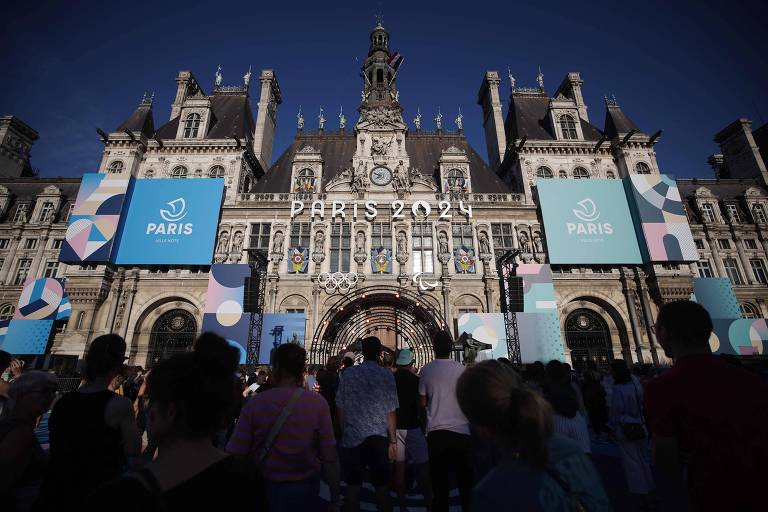 A imagem mostra a fachada de um edifício histórico em Paris, decorado para um evento relacionado aos Jogos Olímpicos de 2024. O edifício é adornado com grandes banners azuis que exibem o texto 'PARIS 2024' e 'PARIS VILLE HÔTE'. Há uma multidão de pessoas na frente, algumas com as costas voltadas para a câmera, e um céu azul claro ao fundo.
