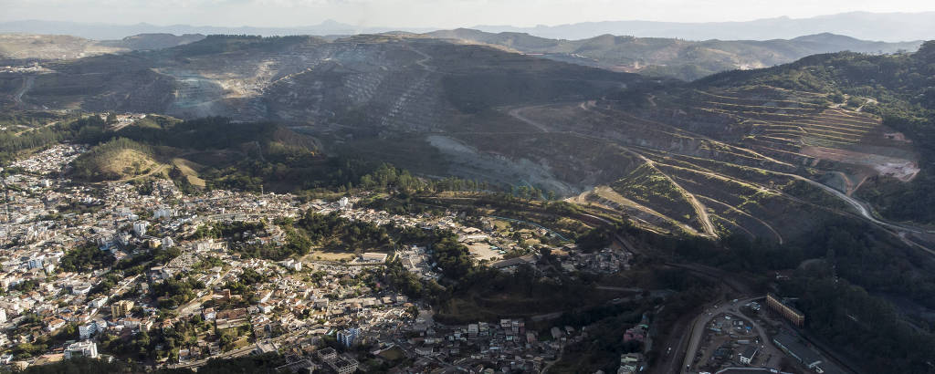 A imagem mostra uma vista aérea de uma cidade situada em uma região montanhosa. A cidade é composta por várias construções e ruas, com áreas verdes visíveis nas encostas das montanhas. Ao fundo, há uma cadeia de montanhas cobertas por vegetação e algumas nuvens no céu. Na parte inferior direita, é possível ver uma área com um campo esportivo.
