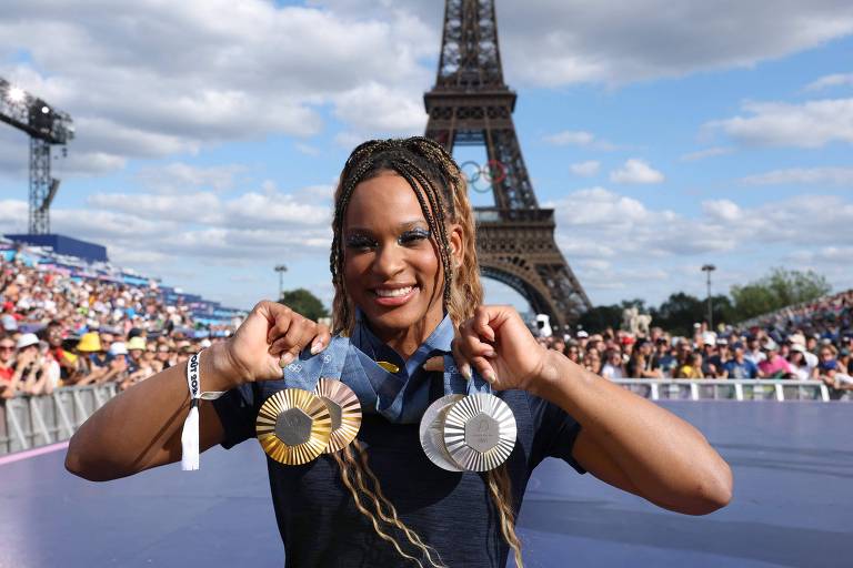 Fotografia mostra a atleta Rebeca Andrade, mulher negra, posando com quatro medalhas, uma de ouro, duas de prata e uma de bronze, em frente à Torre Eiffel. O céu está parcialmente nublado e há uma multidão ao fundo, assistindo ao evento.