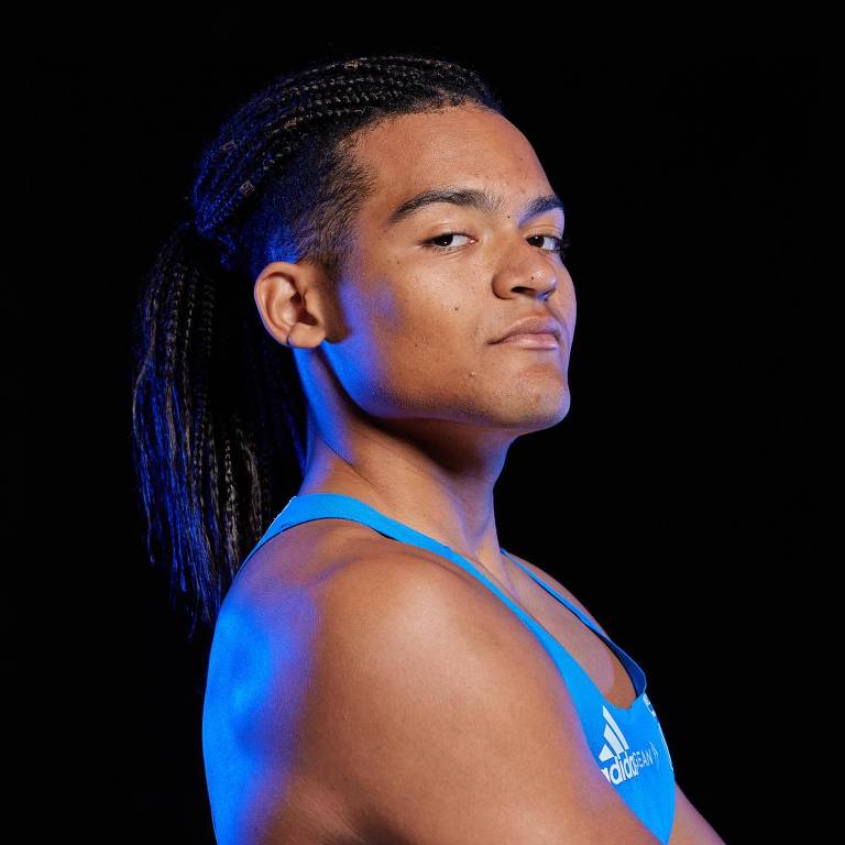 Um atleta com cabelo trançado, usando uma camiseta azul, está posando em um estúdio com fundo preto