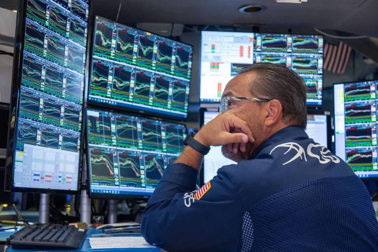 Um homem está sentado em frente a várias telas de computador que exibem gráficos financeiros e dados de mercado. Ele está com a mão no queixo, pensativo, e usa uma camisa azul com detalhes em branco. As telas mostram gráficos coloridos com linhas e barras, indicando flutuações de preços e volumes de negociação.
