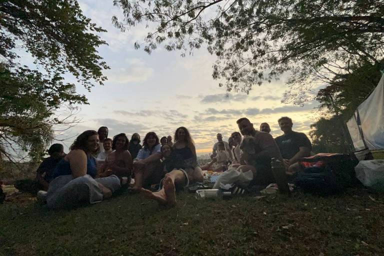 A imagem mostra um grupo de pessoas sentadas em um gramado durante o entardecer. Elas estão dispostas em um círculo, com algumas pessoas sorrindo e outras olhando para a câmera. Ao fundo, há árvores e um céu com nuvens, indicando que o sol está se pondo