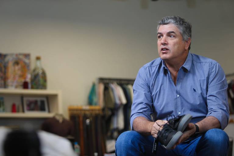 Um homem de cabelos grisalhos, vestindo uma camisa azul clara e jeans, está sentado em um ambiente interno. Ele segura um sapato preto e parece estar em uma conversa ou reflexão. Ao fundo, há prateleiras com roupas e objetos decorativos, além de uma parede clara.