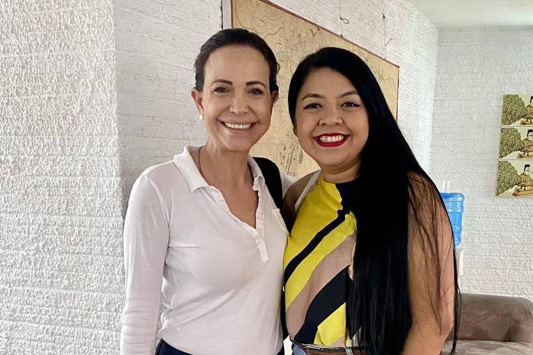 A imagem mostra duas mulheres sorrindo uma para a outra. A mulher à esquerda está vestindo uma camisa branca de manga longa e calças escuras, enquanto a mulher à direita usa uma blusa colorida com listras em amarelo, preto e rosa. O fundo é uma parede clara com um quadro e uma garrafa de água visível