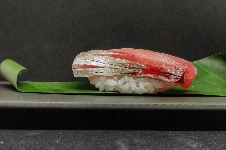 A imagem mostra um sushi colocado sobre uma folha verde em um prato escuro. O sushi é composto por arroz branco moldado e coberto com uma fatia de peixe, que apresenta uma parte prateada e uma parte vermelha. O fundo da imagem é preto, destacando o sushi e a folha.