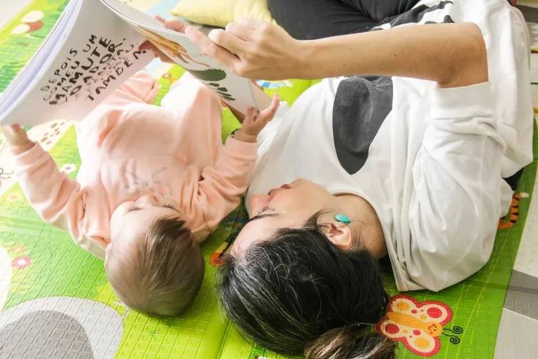 Uma mãe e seu bebê estão deitados em um tapete colorido, lendo um livro. A mãe está segurando o livro com uma mão e a outra mão do bebê está levantada, segurando a página. O bebê está vestindo um macacão rosa e a mãe está usando uma camiseta branca com um desenho. O ambiente é iluminado e acolhedor.