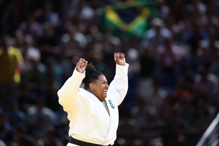 Uma atleta de judô, vestindo um quimono branco, levanta os braços em celebração. Ao fundo, uma multidão está presente, com uma bandeira do Brasil visível. A atleta demonstra alegria e emoção em um evento esportivo.
