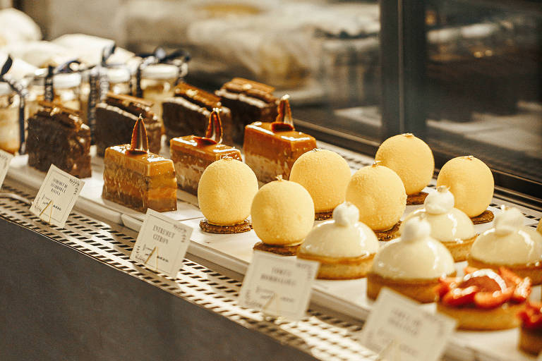 A imagem mostra uma vitrine de doces com uma variedade de sobremesas dispostas em uma prateleira. Há bolos retangulares cobertos com chocolate, doces em formato de ovo amarelo e tortas com cobertura branca. Cada doce tem uma pequena etiqueta com o nome. O fundo é desfocado, sugerindo um ambiente de confeitaria.