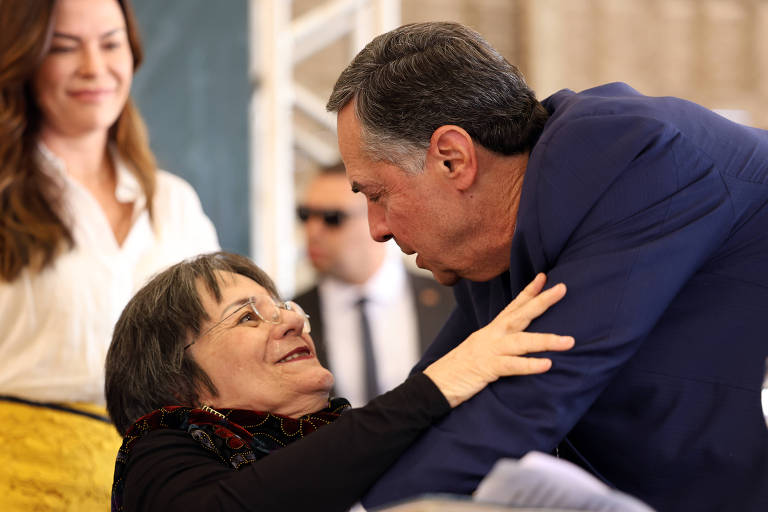 A imagem mostra duas pessoas em um momento de interação. Uma mulher com cabelo curto e grisalho, usando um casaco escuro, sorri enquanto é cumprimentada por um homem de terno azul. Ao fundo, uma mulher com cabelo longo e castanho claro observa a cena. O ambiente parece ser um evento formal, com uma estrutura de fundo que sugere um espaço interno.