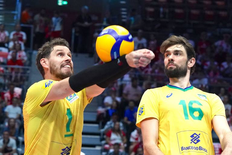 Dois jogadores de vôlei estão em uma quadra. O jogador à esquerda, com a camisa amarela e número 1, está se preparando para fazer um saque, enquanto o jogador à direita, com a camisa amarela e número 16, observa. O ambiente é uma arena esportiva com espectadores ao fundo.