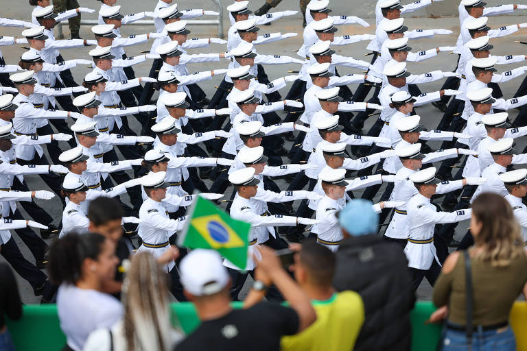 A imagem mostra um grupo de militares em uniforme branco e preto, alinhados em formação, realizando uma apresentação. Eles estão com os braços estendidos, em uma posição de saudação. Na parte inferior da imagem, há um grupo de pessoas assistindo ao desfile, algumas segurando uma bandeira do Brasil. O ambiente parece ser ao ar livre, possivelmente durante um evento cívico.