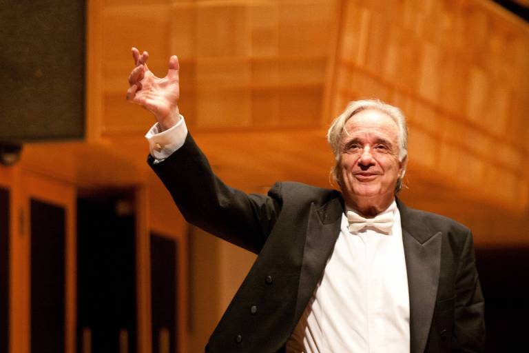 João Carlos Martins, maestro de cabelos grisalhos, vestido com um fraque preto e uma gravata borboleta branca, está em pé em um palco, levantando a mão direita em um gesto expressivo. O fundo apresenta uma arquitetura de auditório com painéis de madeira.
