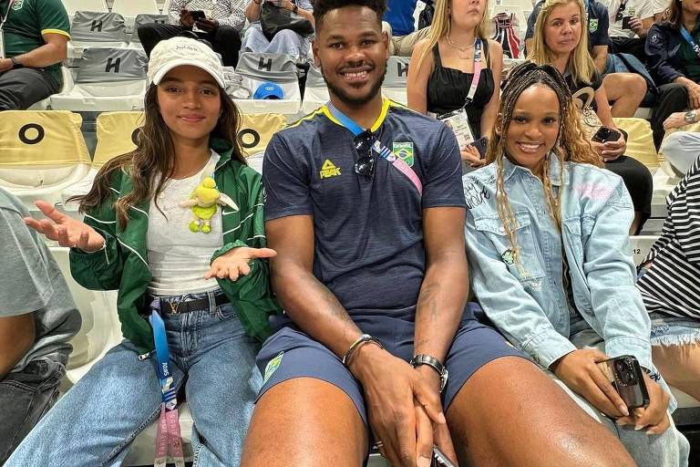 A imagem mostra três pessoas sentadas em um estádio. À esquerda, uma jovem com cabelo longo e solto, vestindo uma jaqueta verde e uma camiseta branca, segura um pequeno boneco verde. No centro, um homem alto e musculoso, usando uma camiseta azul com detalhes em verde e shorts, sorri para a câmera. À direita, uma mulher com cabelo trançado, vestindo uma jaqueta jeans, também sorri. Ao fundo, várias pessoas assistem ao evento, algumas usando camisetas de times e outras com celulares.