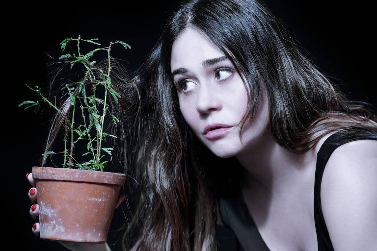 A imagem mostra uma mulher com cabelo longo e solto, olhando para um vaso de planta que ela segura com uma das mãos. A planta parece estar murcha, com ramos finos e folhas pequenas. O fundo é escuro, destacando a expressão preocupada da mulher e o vaso de barro. Ela está vestida com uma blusa preta e tem unhas pintadas de vermelho.