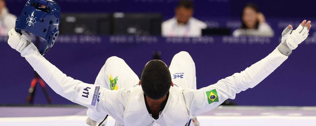 Um atleta de esgrima está deitado no chão, com os braços abertos e a cabeça voltada para cima. Ele usa um uniforme branco e um capacete azul. Ao fundo, há pessoas assistindo ao evento em um ambiente esportivo.