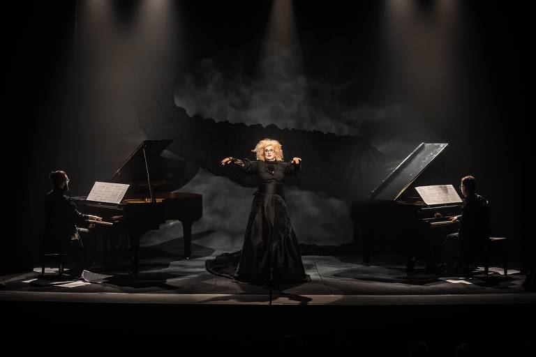 A imagem mostra uma performance em um palco iluminado. No centro, uma artista vestida com um vestido longo e escuro está em pé, com os braços levantados, enquanto dois pianistas, um à esquerda e outro à direita, tocam pianos de cauda.