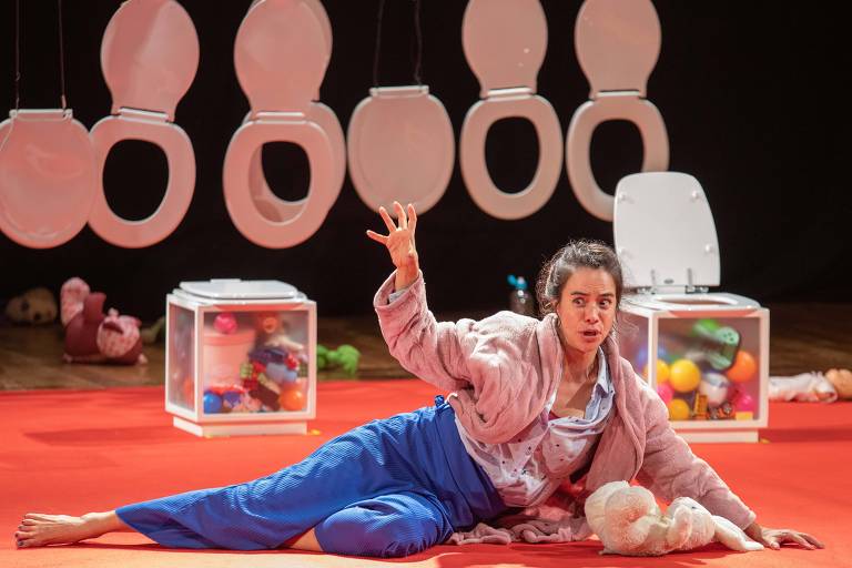 A imagem mostra uma atriz em uma performance teatral, vestindo um roupão rosa e calças azuis. Ela está deitada no chão vermelho, com uma expressão facial intensa, gesticulando com a mão direita.