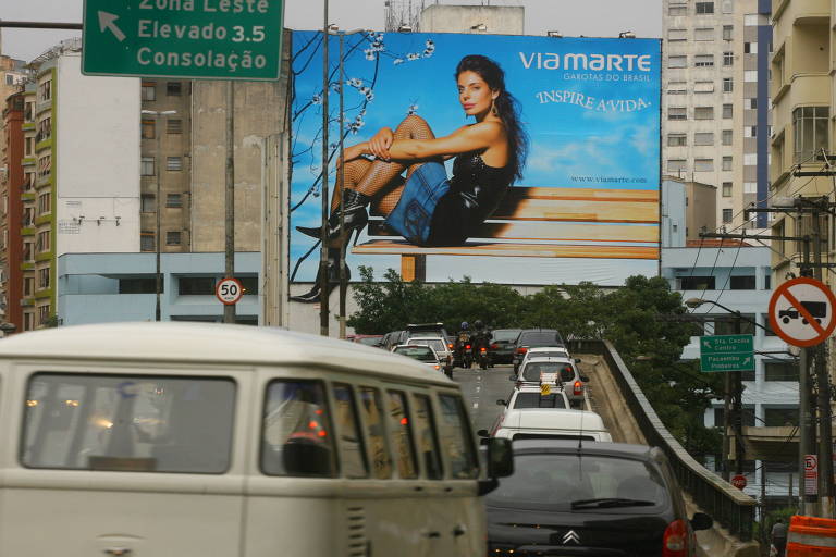 A imagem mostra um outdoor publicitário em São Paulo, com uma mulher sentada em um banco, vestindo uma blusa preta e calças jeans. O outdoor é da marca Viamarte e contém a frase 'INSPIRE A VIDA'. Ao fundo, há prédios urbanos e um tráfego de veículos na via em primeiro plano. Placas de sinalização indicam direções e limites de velocidade. 