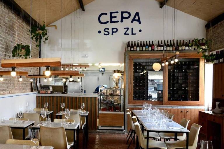 A imagem mostra o interior de um restaurante chamado CEPA. O ambiente possui mesas com toalhas brancas e cadeiras de plástico amarelo. As paredes são de tijolos expostos e há plantas suspensas no teto. No fundo, há uma área de cozinha visível e uma prateleira com garrafas de vinho. A iluminação é suave, com lâmpadas penduradas. O nome 'CEPA' está escrito em letras grandes na parede.