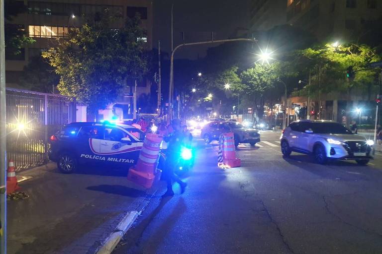 A imagem mostra uma cena noturna em uma rua, onde um carro da Polícia Militar, com luzes azuis e vermelhas piscando, está estacionado. Há cones de sinalização laranja ao redor, e alguns policiais estão visíveis. Vários veículos estão passando pela rua, e há iluminação de postes e janelas de prédios ao fundo