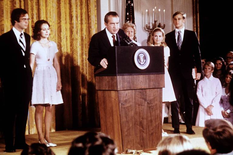 A imagem mostra um evento formal em que Richard Nixon está falando ao microfone em um pódio com o selo presidencial. Ele está cercado por quatro pessoas: duas mulheres e dois homens, todos vestidos de maneira formal. Ao fundo, há uma cortina dourada e uma bandeira dos Estados Unidos. O público está visível na parte inferior da imagem, assistindo ao discurso.