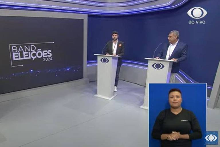 A imagem mostra um estúdio de televisão com três pessoas em pé atrás de pódios. No centro, um apresentador fala, enquanto dois candidatos estão à sua esquerda e direita. Ao lado direito da imagem, há uma intérprete de libras. O fundo é escuro com o texto 'Rádio Eleições' em destaque.
