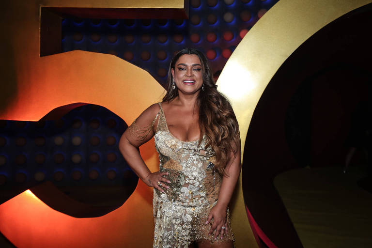 A imagem mostra uma mulher sorridente em um evento, vestindo um vestido brilhante e justo, com detalhes em dourado e prateado. Ela está posando em frente a um fundo com números grandes em dourado e um padrão de bolinhas em azul. A mulher tem cabelo longo e solto, e usa brincos. O ambiente é festivo e bem iluminado.