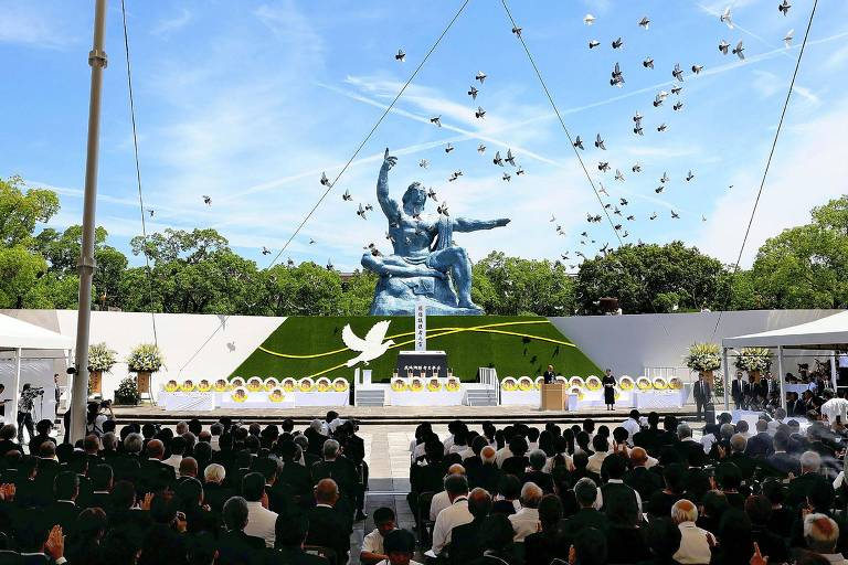 A imagem mostra uma cerimônia ao ar livre com uma grande estátua no fundo, representando uma figura humana em uma pose dinâmica. A estátua é cercada por um gramado verde e há uma estrutura de palco com um altar decorado. O público, vestido de preto, está sentado em cadeiras, observando a cerimônia. No céu, várias pombas estão voando, simbolizando paz. O ambiente é iluminado por um céu azul claro com algumas nuvens.