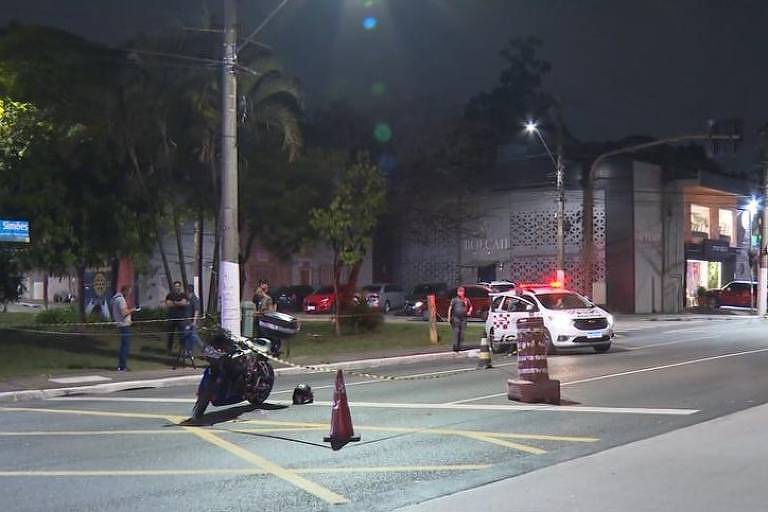 imagem mostra, em primeiro plano, motocicleta isolada por cones de trânsito. ao fundo, uma viatura da polícia, com sirenes ligadas. ao lado esquerdo, alguns homens observam a cena. 