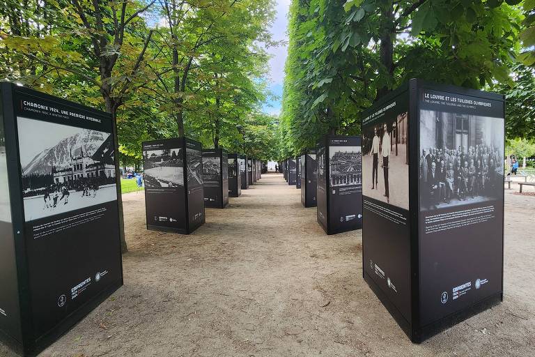 A imagem mostra uma exposição ao ar livre em um parque, com painéis informativos dispostos em fileiras ao longo de um caminho de terra. Os painéis são de cor preta e apresentam fotografias e textos, com árvores verdes ao redor. O céu está parcialmente nublado, com algumas áreas de azul visíveis.