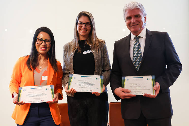 Três pessoas estão em pé， segurando certificados em uma cerimônia de premiação. A primeira pessoa à esquerda usa uma blusa preta e um blazer laranja， a segunda pessoa no meio usa uma blusa preta e tem cabelo longo e solto， e a terceira pessoa à direita usa um terno escuro e uma gravata com padrões. Todos estão sorrindo para a câmera.