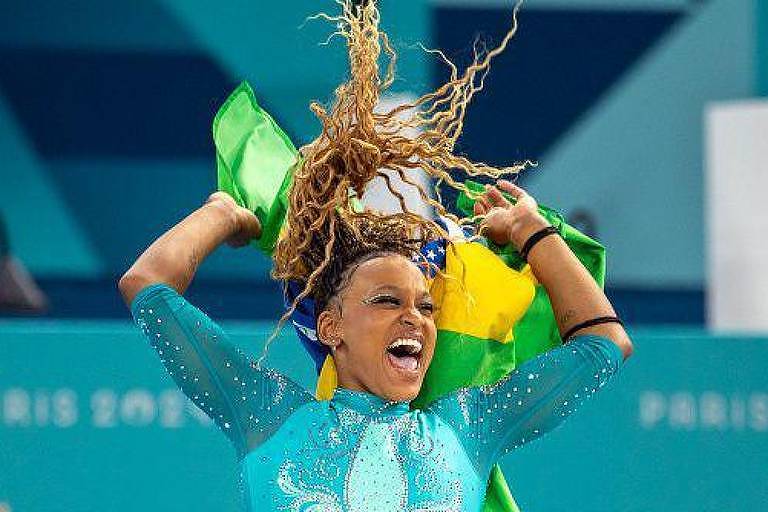 Uma atleta está em uma competição, vestindo um traje azul com detalhes brilhantes. Ela está sorrindo e levantando a bandeira do Brasil, que é verde e amarelo, acima da cabeça. Seu cabelo está solto e se movimenta com a ação. Ao fundo, há uma parede com a inscrição 'PARIS 2024'.
