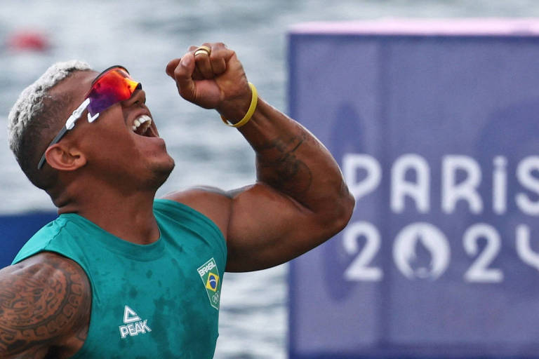 Um atleta brasileiro, com cabelo platinado e óculos de sol, está comemorando com um punho cerrado e um sorriso largo. Ele usa uma camiseta verde com a bandeira do Brasil e está em um ambiente aquático, com um fundo desfocado que mostra a água e um painel com a inscrição 'PARIS 2024'.
