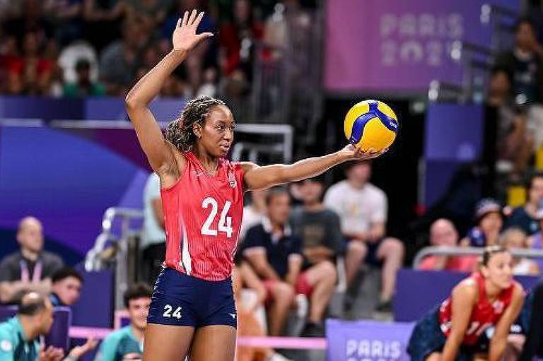 Uma jogadora de vôlei está se preparando para sacar. Ela usa uma camiseta vermelha com o número 24 e shorts pretos. A jogadora levanta a mão esquerda enquanto segura uma bola de vôlei amarela e azul com a mão direita. Ao fundo, é possível ver uma plateia e outros jogadores em um ambiente de competição.
