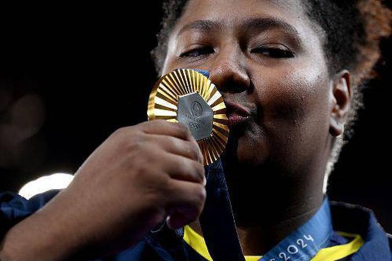 A imagem mostra uma atleta segurando uma medalha de ouro perto do rosto, enquanto a beija. Ela tem cabelo crespo e usa uma jaqueta azul com detalhes em amarelo. O fundo é desfocado, destacando a atleta e a medalha.
