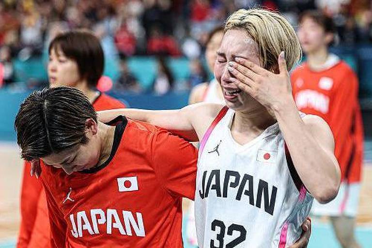 A imagem mostra duas jogadoras de basquete do Japão em um momento emocional após uma partida. Uma delas, vestindo uma camisa branca com o número 32, está chorando e se enxugando as lágrimas, enquanto a outra, com uma camisa vermelha, a apoia, inclinando a cabeça para baixo. Ao fundo, outras jogadoras e torcedores são visíveis, criando um ambiente de tensão e emoção.
