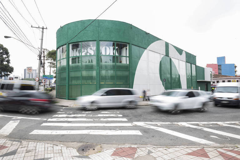 Fachada da loja de móveis situada em uma esquina. A loja é verde escura, com detalhes em branco. Carros passam na rua em frente à fachada e o céu está nublado.