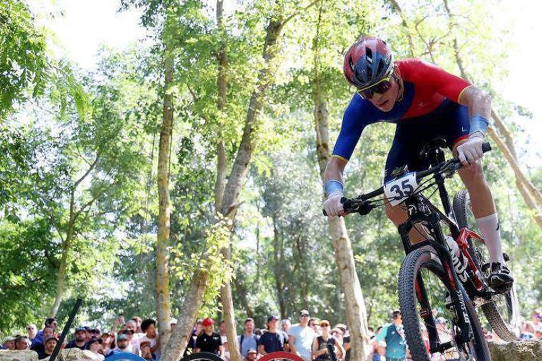 Um ciclista em uma competição de mountain bike, usando um uniforme azul e vermelho, está saltando sobre uma pedra em um percurso ao ar livre. Ao fundo, há uma multidão assistindo, com árvores verdes ao redor.
