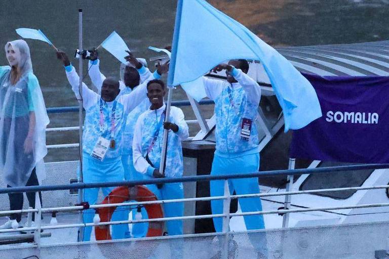 A imagem mostra um grupo de pessoas em um barco, celebrando com bandeiras azuis. Uma mulher está à esquerda, vestindo uma capa de chuva transparente, enquanto os homens à direita estão vestidos com roupas azuis e seguram bandeiras. Ao fundo, há uma bandeira roxa com a palavra 'SOMALIA' escrita em branco. O ambiente parece ser ao ar livre, possivelmente em um evento ou competição.
