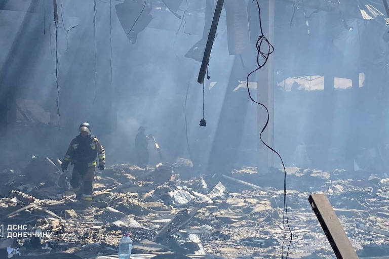 A imagem mostra um ambiente devastado, com escombros e fumaça. Dois bombeiros estão visíveis, um deles está mais próximo, vestindo equipamento de proteção. O local parece ter sofrido um incêndio significativo, com destroços espalhados pelo chão e luzes que penetram através da fumaça.