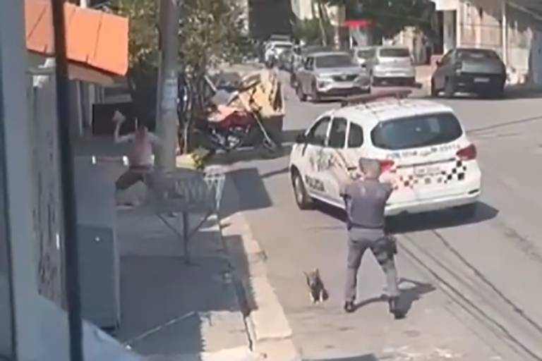 reprodução de vídeo mostra policial de costas apontando arma para homem que se prepara para jogar tijolo contra ele. há uma viatura à frente do policial, que está a cerca de três metros de distância do homem, um cachorro caminha perto do policial