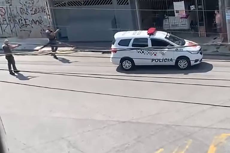 reprodução de vídeo mostra dois policais ao lado esquerdo apontando suas armas para homem do lado direito da imagem, em frente a um portão há uma viatura à frente dos policiais, que estão a cerca de quatro metros do homem