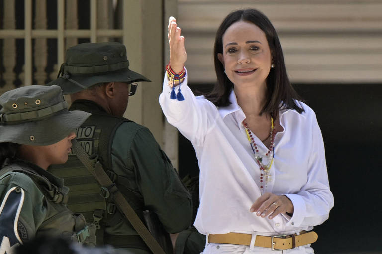 A imagem mostra uma mulher vestindo uma camisa branca e sorrindo, acenando com a mão direita. Ela está em primeiro plano, enquanto ao fundo há dois homens em uniformes militares, um deles usando um boné. A mulher usa acessórios coloridos e parece estar em um ambiente ao ar livre.