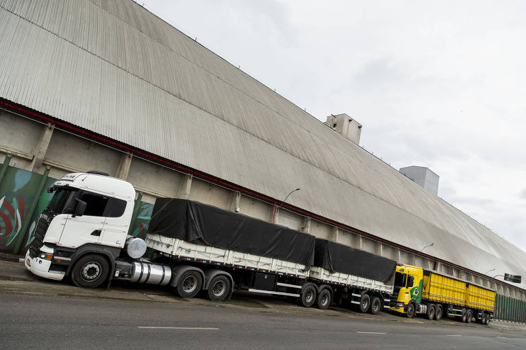 A imagem mostra dois caminhões estacionados em frente a um grande edifício de concreto. O caminhão da esquerda é branco com uma carroceria preta coberta por uma lona, enquanto o caminhão da direita é amarelo. O céu está nublado, e a estrutura ao fundo é alta e retangular