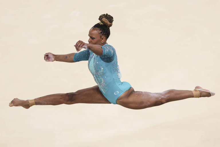 Rebeca, mulher negra, está de collant verde-água de mangas compridas e coque no alto da cabeça. Ela faz uma acrobacia no ar, com as pernas abertas. O tablado é bege