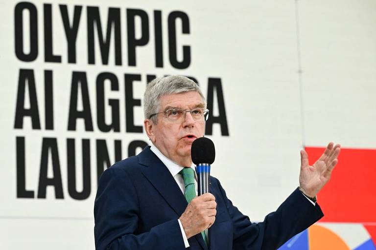 Thomas Bach, presidente do COI, é um homem de cabelo grisalho, usando um terno escuro e uma gravata, está falando ao microfone. Ao fundo, há um grande texto em letras maiúsculas que diz 'OLYMPIC AI AGENDA LAUNCH'. O ambiente parece ser um evento formal.